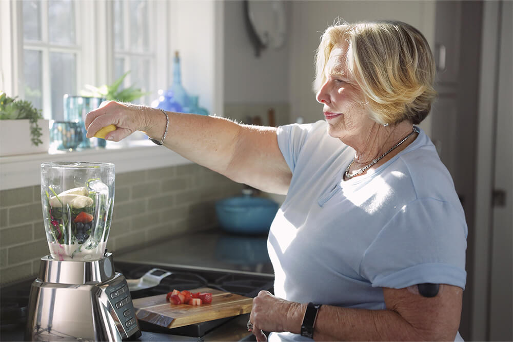 Mujer haciendo un batido de frutas mientras lleva puesto un dispositivo de monitorización continua de la glucosa.
